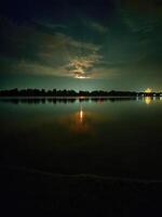 Color photography of evening nature by the river. Rising full moon above the trees. Beautiful reflection of the moon path in the water. photo