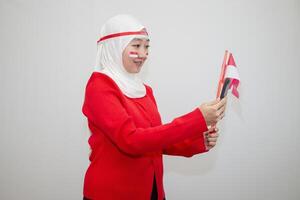 Woman in white hijab wearing red sweater. celebrating Indonesia's independence day. holding a flag and taking a selfie with a cellphone. isolation on white background. photo