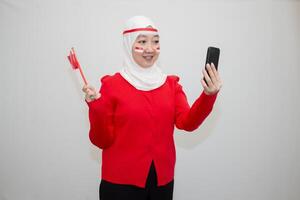 Woman in white hijab wearing red sweater. celebrating Indonesia's independence day. holding a flag and taking a selfie with a cellphone. isolation on white background. photo