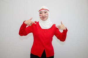 Woman in white hijab wearing red sweater. celebrating Indonesia's independence day. pointing downwards. isolation on white background. photo