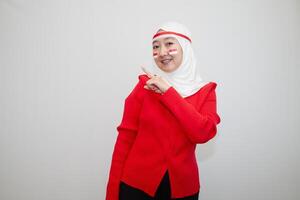 Woman in white hijab wearing red sweater. celebrating Indonesia's independence day. pointing to the side. isolation on white background. photo