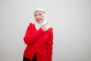 Woman in white hijab wearing red sweater. celebrating Indonesia's independence day. pointing to the side. isolation on white background. photo
