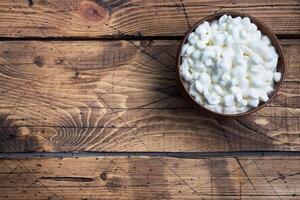 Fresh cottage cheese grain in a wooden bowl. Curd in granules with cream. Wooden rustic background. Copy space. photo
