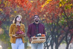 Happy caucasian farmer couple carrying organics homegrown produce harvest with apple, squash and pumpkin while walking along the country road with fall color from maple tree during autumn season photo