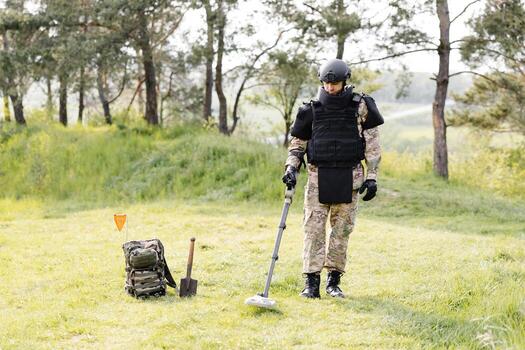 A man in a military uniform and bulletproof vest works in the forest with a metal detector. A minesweeper performs work on demining the territory photo