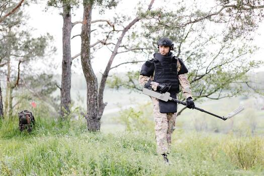 A man in a military uniform and bulletproof vest works in the forest with a metal detector. A minesweeper performs work on demining the territory photo