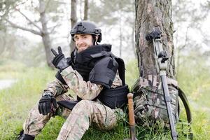 A man in a military uniform and a bulletproof vest sits in the forest near a metal detector and a military backpack. A man pauses in the work of demining the territory photo
