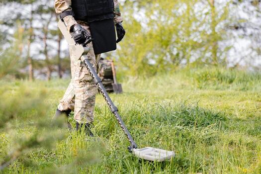 A man in a military uniform and bulletproof vest works in the forest with a metal detector. A minesweeper performs work on demining the territory photo