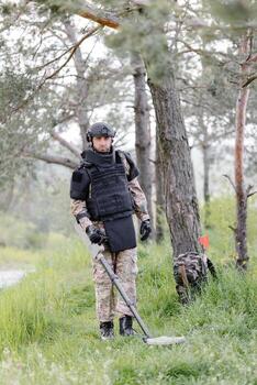 A man in a military uniform and bulletproof vest works in the forest with a metal detector. A minesweeper performs work on demining the territory photo