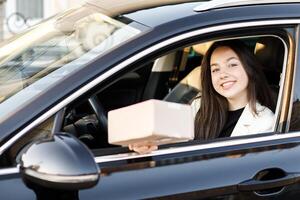 Young and happy business woman giving a parcel through the vehicle window on the go while driving a car. Concept of fast delivery on the go photo