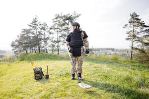 A man in a military uniform and bulletproof vest works in the forest with a metal detector. A minesweeper performs work on demining the territory photo
