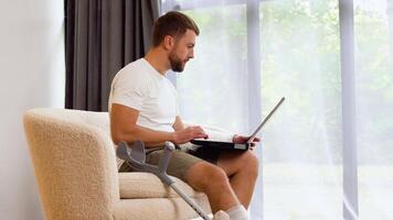 Handsome man with broken leg and arm in gypsum is using a laptop and smiling while sitting on sofa at home video