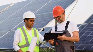 Two diverse workers engineers talking with each other while holding a technical documentation with a large solar panel in background video