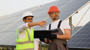 Two diverse workers engineers talking with each other while holding a technical documentation with a large solar panel in background video