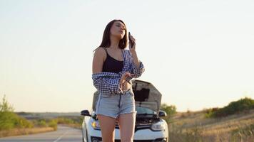 Upset young woman driver talking angrily on phone with assistance service near a broken car with open hood while inspecting engine having trouble with her vehicle video