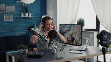 Creative digital content creator putting on headphones and starting to record while talking to audience. Friendly young woman streaming live and remote podcast show while sitting in studio. video