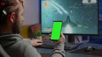 Concentrate gamer holding phone with green screen mock up chorma key display while playing games on powerful computer streaming online competition. Player using isolated desktop playing shooter games video