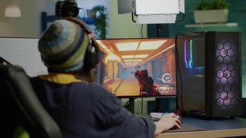 Back shot of pro streamer with headphone playing FPS videogame during esport competition using RGB powerful computer. Player sitting on gaming chair using streaming equipment late night in home studio video