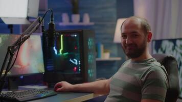 Streamer man looking at camera smiling while streaming videogames using stream chat. Pro cyber videogamer playing space shooter game at RGB powerful personal computer in gaming home studio video