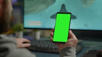 Competitive gamer looking at smartphone with green screen chroma key desktop playing online games on powerful computer during live tournament. Cyber player using phone with mock up isolated display video
