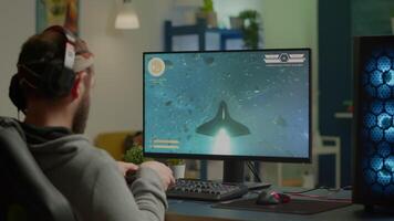 Pro man gamer playing game at powerful personal computer late night wearing professional headphones. Excited player using wireless controller for virtual tournament gaming space shooter at home video
