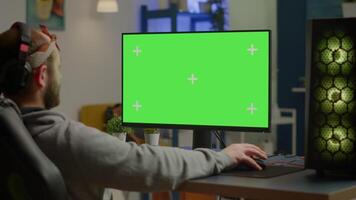 Gamer playing games on powerful computer with green screen chroma key desktop mock-up display in gaming home studio. Player using RGB keyboard with isolated monitor streaming game wearing headset video