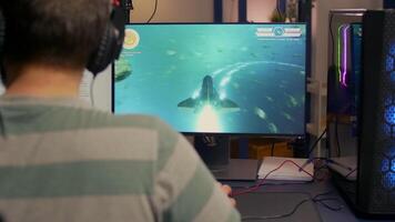 Over shoulder footage of professional streamer playing digital space shooter games on computer using headphones, microphone and mouse. Videogamer man streaming online games in room with RGB video