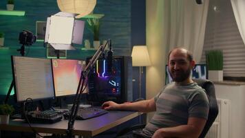 Streamer man looking at camera and smiling while streaming chat is open. Videogamer streamer playing space shooter game at gaming home studio equipped with professional equipment video