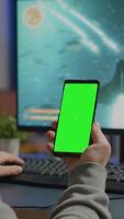 Vertical . Concentrate gamer holding phone with green screen mock up chorma key display while playing games on powerful computer streaming online competition. Player using isolated desktop playing shooter games video