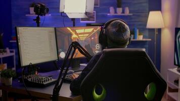 Back shot of pro streamer with headphone playing first person shooter game competition use RGB keypad. Player sitting on gaming chair using streaming equipment late at night in home studio video