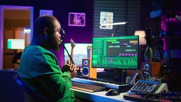 African american sound engineer using midi controller notes to add audio effects in post production, editing tracks with digital software and mixing console. Male musician in home studio. Camera B. video