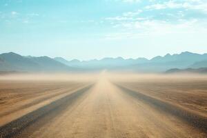 A long, deserted gravel road stretches into the horizon, surrounded by a parched landscape and distant mountains under a bright blue sky, creating a scene of tranquility and inviting exploration photo