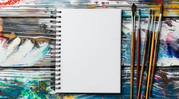 A blank sketchbook is thoughtfully positioned on a colorful, paintsplattered surface adorned with an array of brushes, making it an ideal tool for nurturing art and design inspiration in all forms photo
