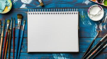 An empty sketchbook lies open, surrounded by various paintbrushes and an array of vibrant paints, creating an ideal environment for artists who wish to unleash their full creativity and imagination photo