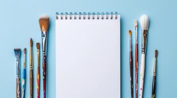 This vibrant mockup showcases various paint brushes neatly arranged with a blank sketchbook on a soothing light blue background, perfect for inspiring artists and showcasing art projects photo