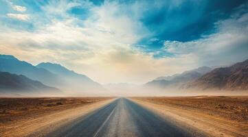 A deserted gravel road stretches endlessly into the distant horizon, surrounded by arid and dry terrain as well as majestic mountains that rise dramatically under a stunning and atmospheric sky photo