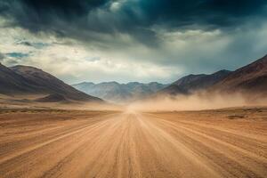 A long gravel road extends towards the distant horizon, bordered by parched, dusty terrain and towering mountains beneath an overcast, moody sky that evokes a sense of adventure and exploration photo
