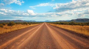 A secluded gravel road extends endlessly towards the distant horizon, enveloped by an arid and barren landscape under a bright blue sky adorned with scattered, fluffy clouds drifting lazily by photo