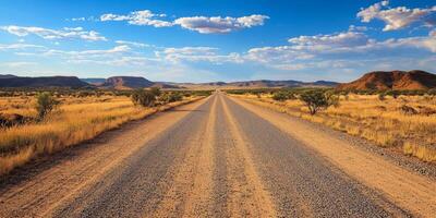 A long and deserted gravel road extends endlessly into the distant horizon, embraced by an arid landscape that unfurls beneath a bright blue sky, adorned with scattered clouds, creating a scenic view photo