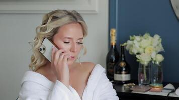 Bride in a robe talking on the phone before the wedding, A candid shot of a bride in a white robe, talking on the phone while sitting in a beautifully decorated room. video