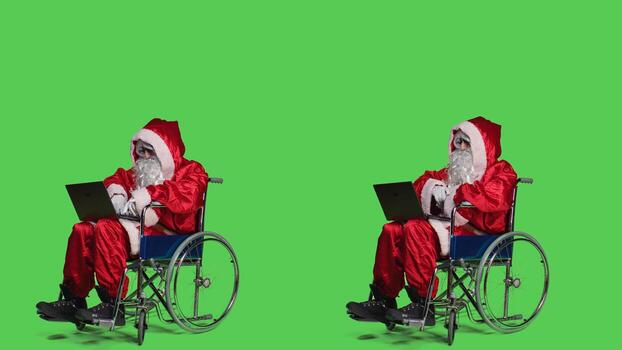 Saint nick with disability using laptop in studio, attending online videocall meeting and browsing webpage. Modern santa character in wheelchair working with internet on portable computer. photo