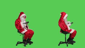 Modern santa works on digital tablet during christmas holiday celebration, looking on online webpage while he sits on chair. Young man against full body greenscreen backdrop using device. photo