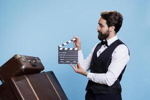 Young bellboy using clapperboard in studio, pretending to work in post production movie filming industry. Hotel porter in formal attire clapping filmslate accessory, saying action on set. photo