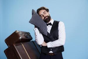 Tired bellboy posing with pillow on camera, expressing burnout and feeling sleepy at professional classy job. Hotel concierge taking quick nap after being overworked, exhausted doorman. photo