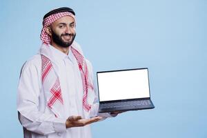Smiling muslim man holding laptop and pointing with hand at empty white screen portrait. Cheerful arab person wearing traditional clothes presenting computer display mockup photo