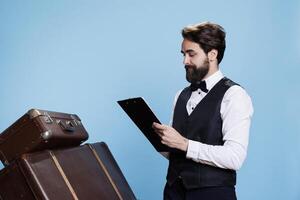 Bellhop reading checklist of tourists, holding catalogue register on clipboard to verify list of guests at hotel. Doorkeeper checking reservations on official log archive papers, take notes. photo
