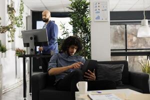 Start up entrepreneur checking project presentation on digital tablet in office. Young arab businessman sitting on couch and using portable gadget to send message in social media photo