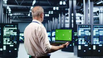 Knowledgeable IT professional navigating network of server racks in industrial hub. Repairman using green screen laptop, ensuring sound operations, doing optimizations and maintenance in data center photo
