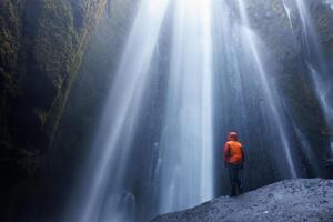 Traveller at seljalandsfoss cascade posing under water stream, river raining over tourists inside of icelandic waterfall. Frozen cold flow near mountain tops and cliffs, majestic scenery. photo