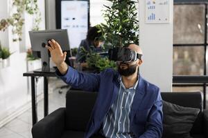 Entrepreneur using vr glasses for market research and analysis while working in business office. Project manager wearing virtual reality headset while sitting on couch in coworking space photo